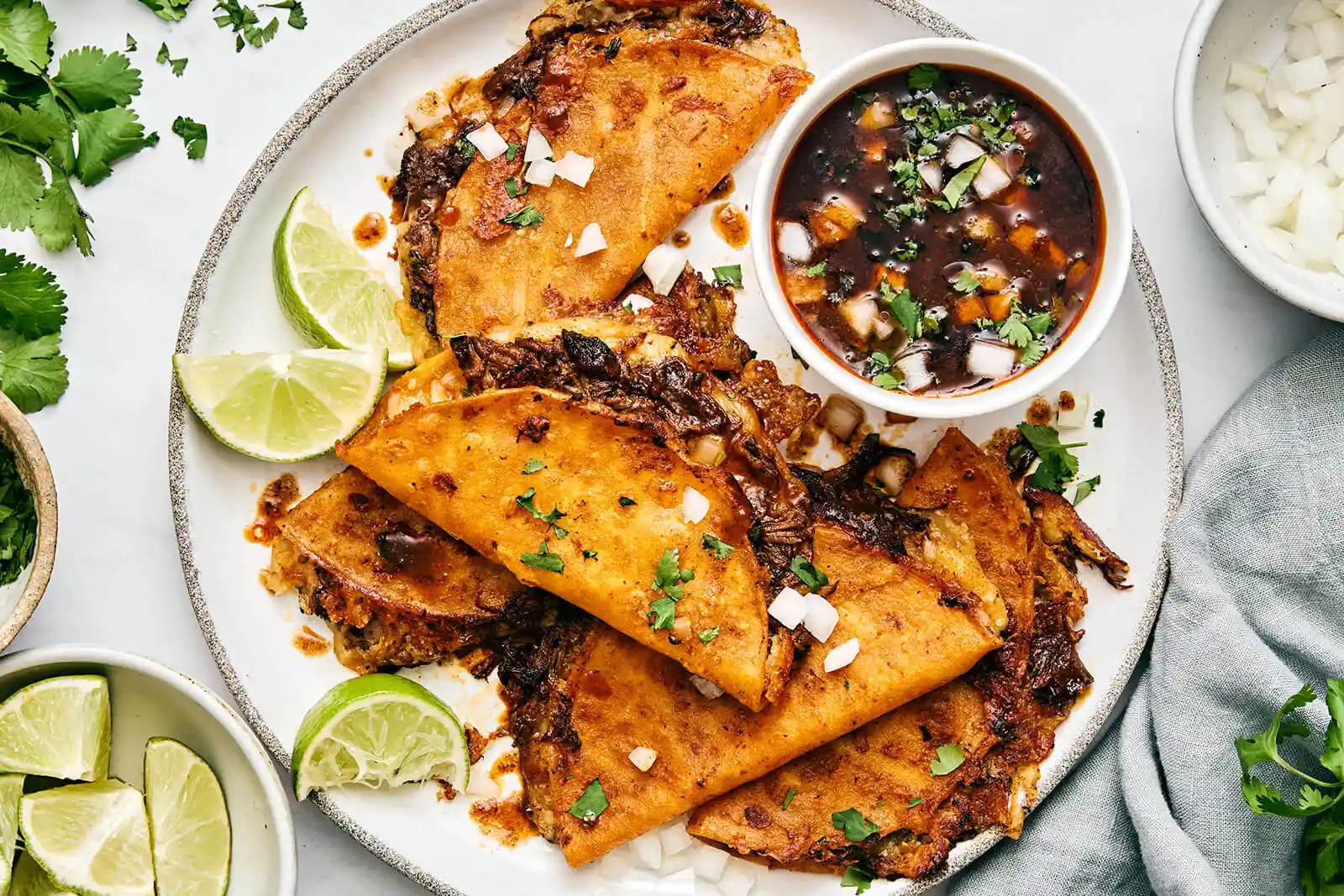 The Ultimate Slow Cooker Beef Birria Tacos (Quesabirria Recipe) 2 Crispy beef birria tacos on a platter with cilantro, onions, and a side of rich consommé dipping sauce